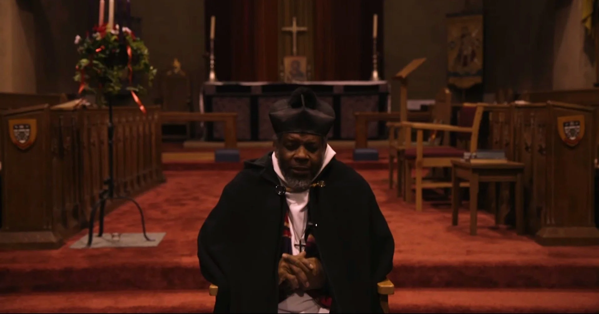 Man kneeling with head bowed and hands clasped in a church with a cross, candles, and chairs in the background.