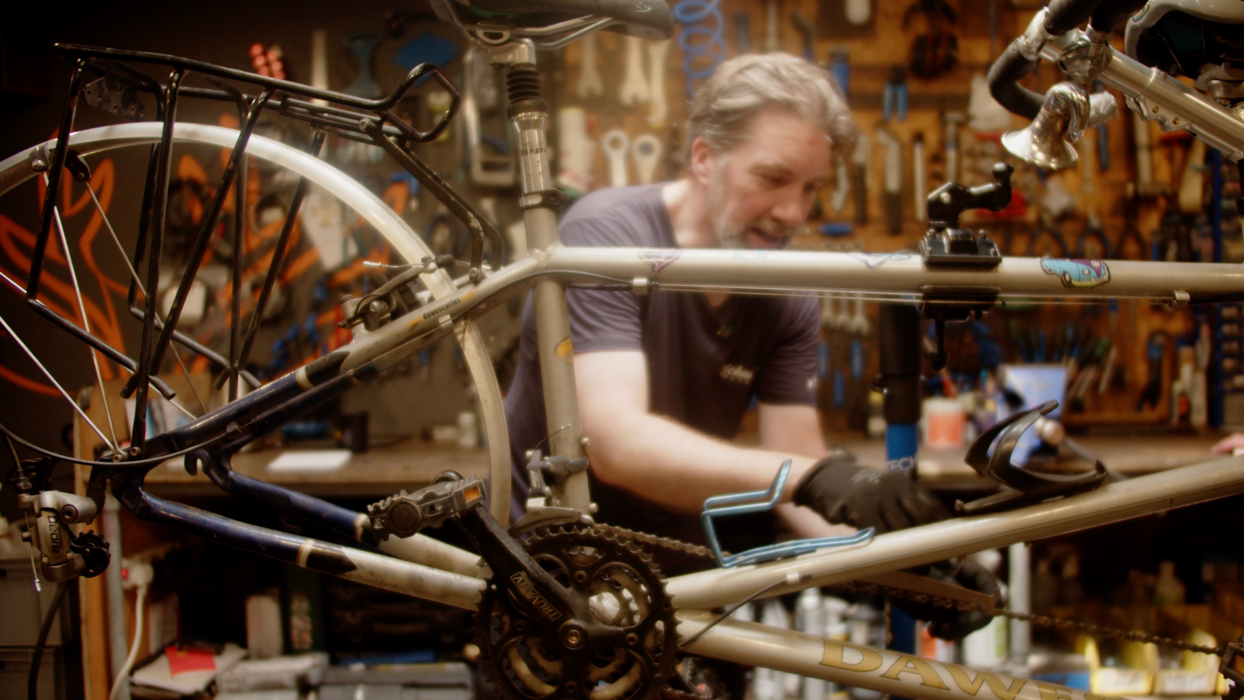 A man working on a bicycle in a workshop, surrounded by tools and parts hanging on the wall.