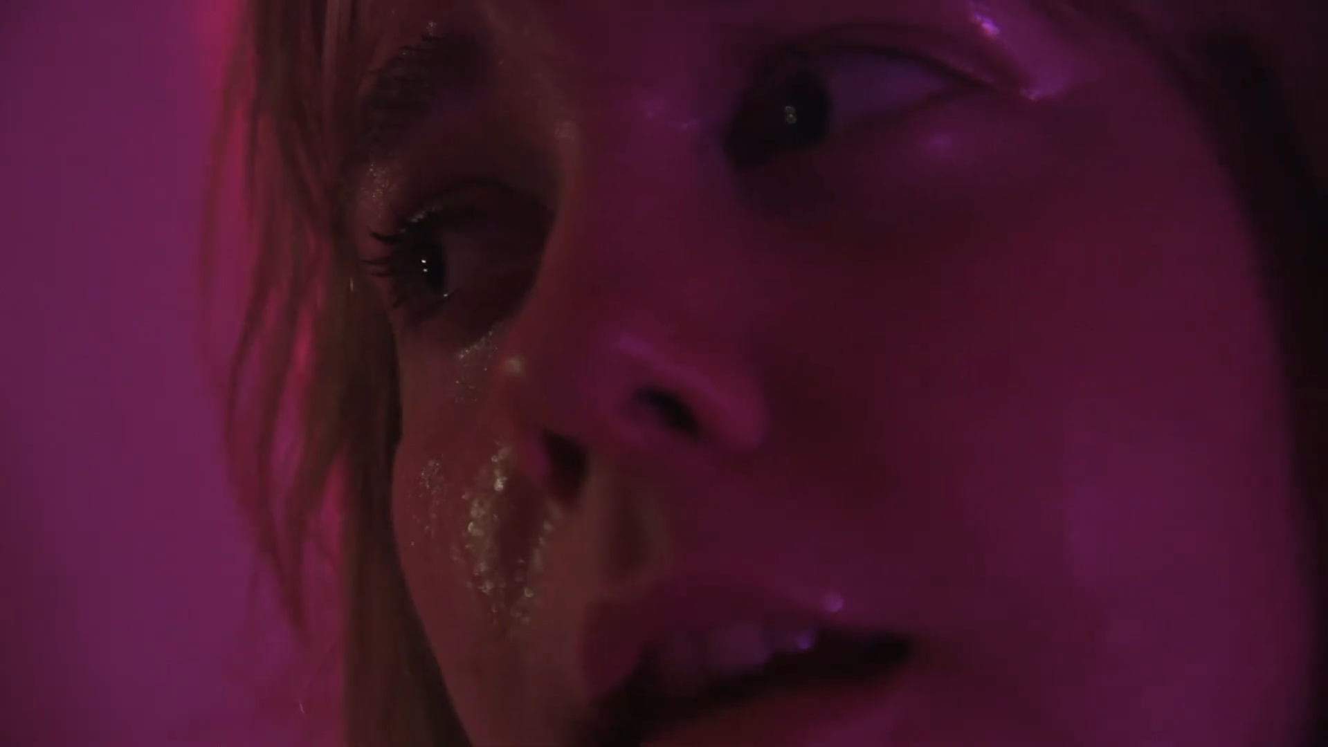 Close-up of a person's face with wet hair and shiny skin, under pink lighting.