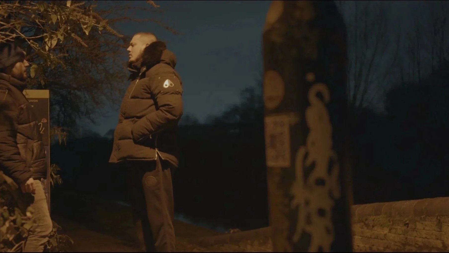 Two men standing and talking outdoors at night, wearing winter jackets. One man has his hands in his pockets, and the other is facing him. The background includes trees and a decorative metal sculpture, with dim lighting.