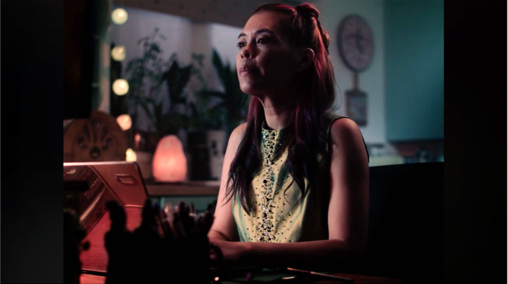 Young woman sitting at a desk in a dimly lit room, illuminated by soft, colorful lighting with plants and decorative items in the background.
