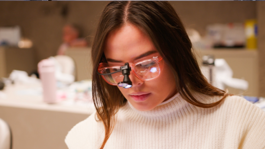 A woman wearing protective glasses with a headlamp is working on a project in a workspace, with various tools and objects blurred in the background.