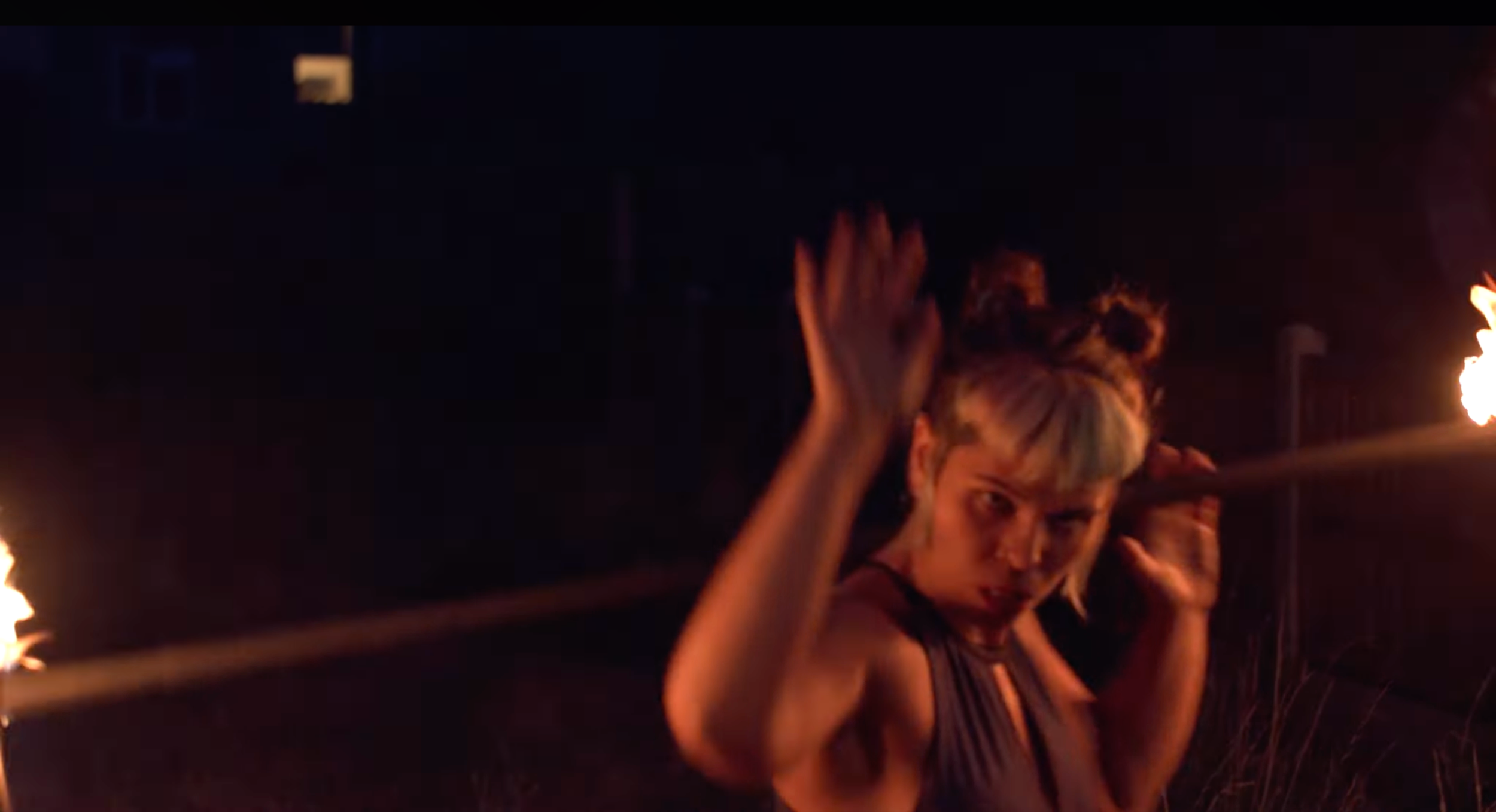 A person with short, light-colored hair holding a flaming torch in a dark environment, with their face partially visible and making a gesture with their hand.