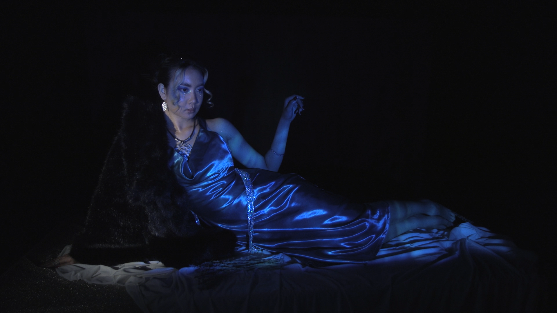 Woman lying on the floor wearing a silky blue dress, illuminated with blue light, with makeup and jewelry, against a black background.