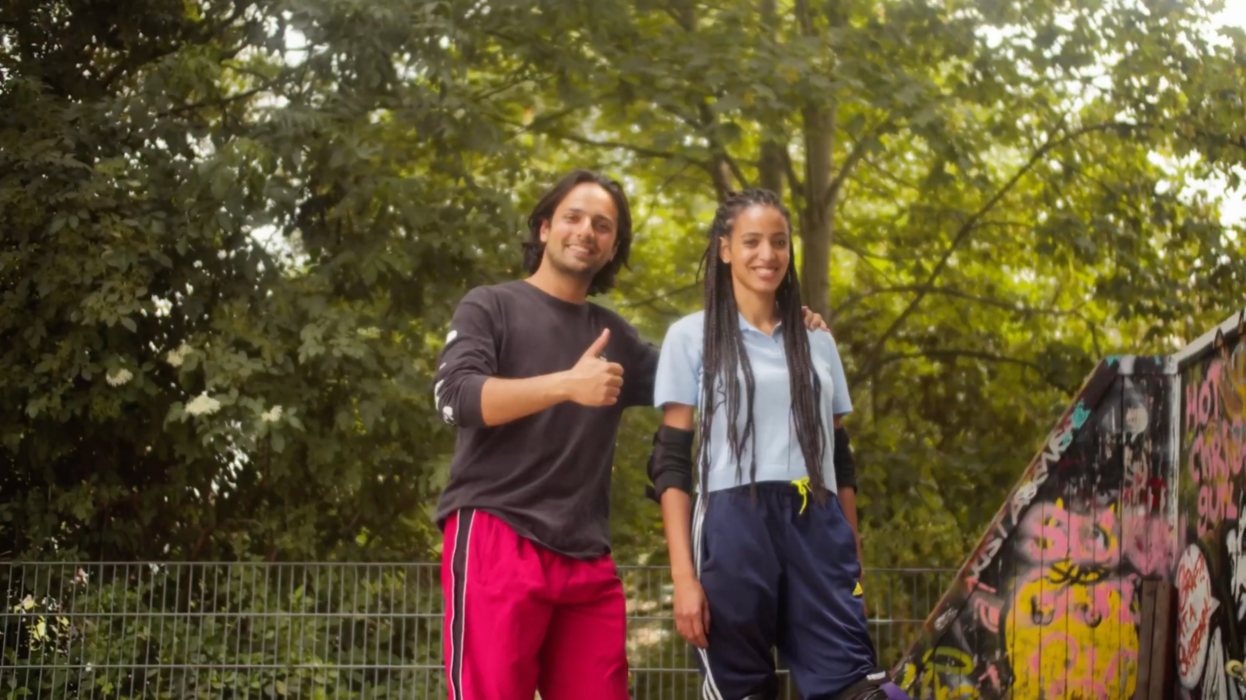 Two young people, a man and a woman, standing outdoors in front of trees, smiling, with the man giving a thumbs-up gesture.