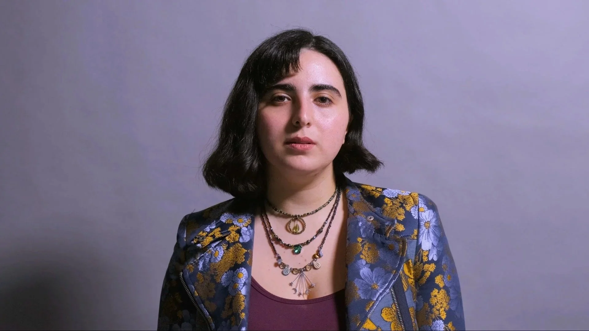 Portrait of a woman with shoulder-length dark hair wearing a blue floral jacket, layered necklaces, and a maroon top, standing against a plain gray background.
