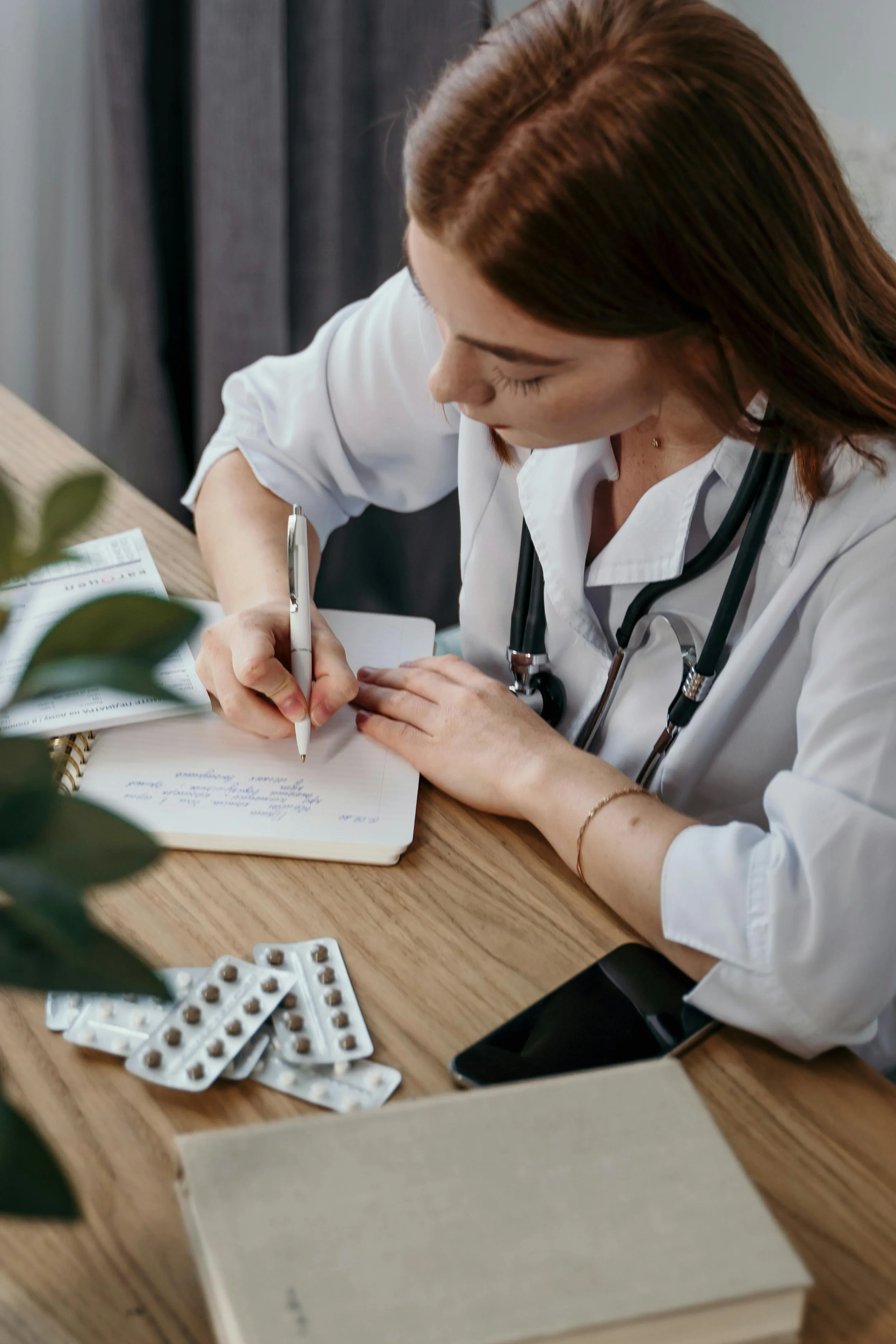 A female doctor with red hair taking notes at her desk, with medication, a smartphone, and a notebook on the desk.