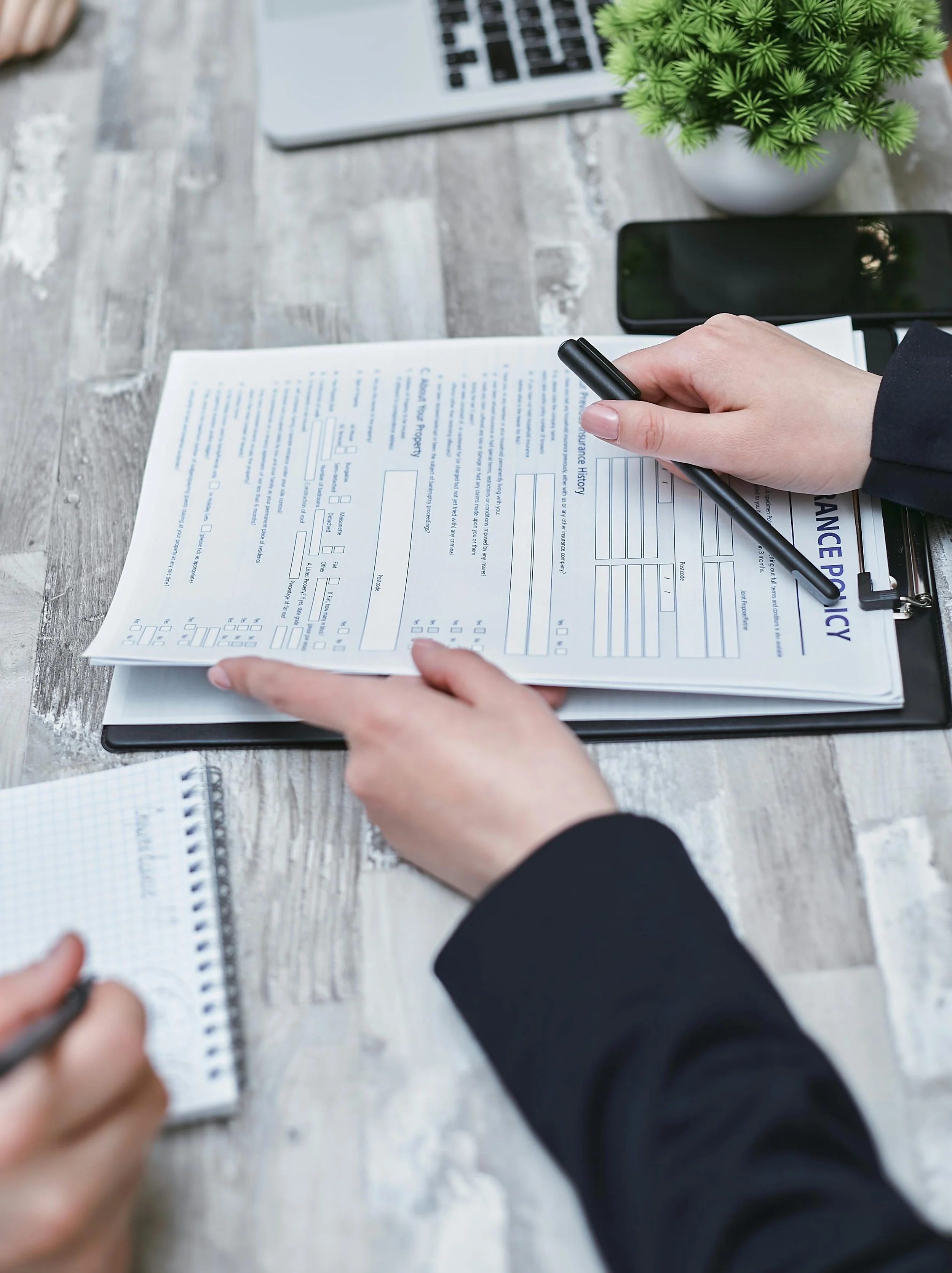Person filling out an insurance form on a clipboard in an office setting, with a potted plant, a smartphone, and a notebook on the desk.