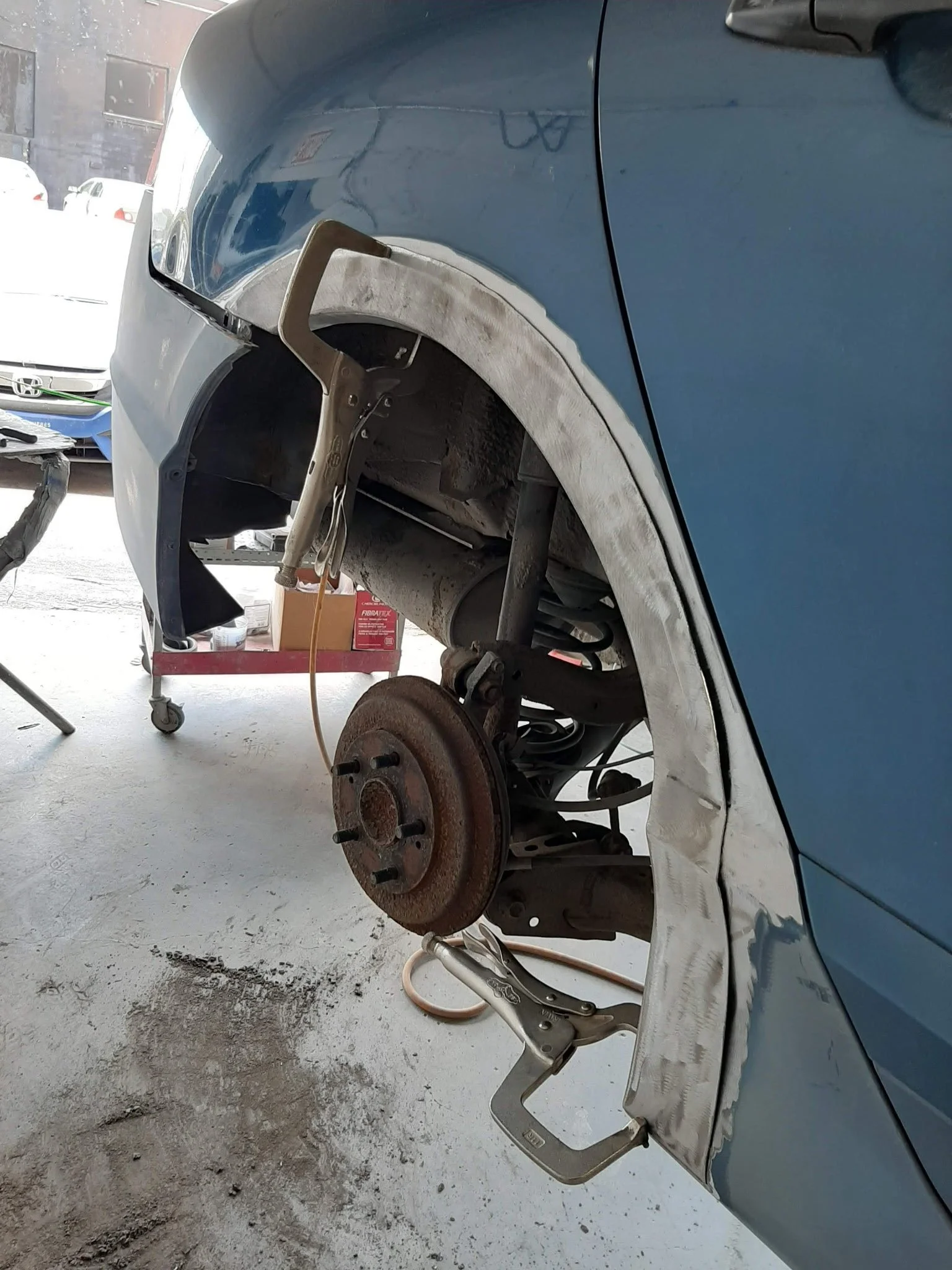 Side view of a car with the front wheel removed, showing the brake rotor, suspension, and other components. The car is elevated and being repaired in a garage.