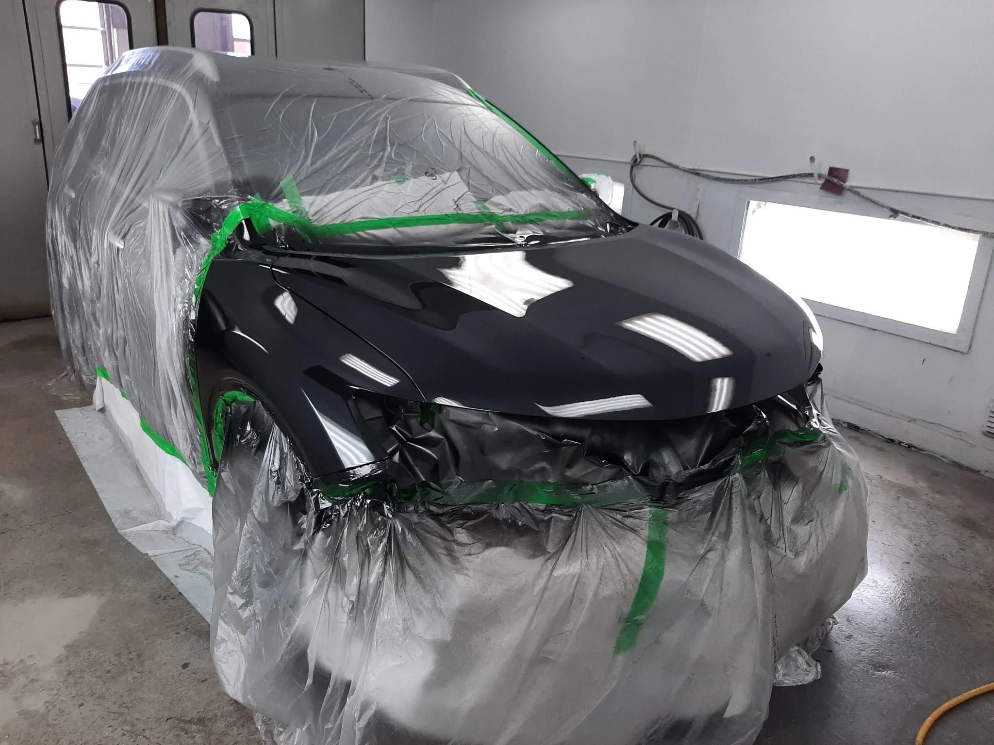 Black sports car in a paint booth, partially covered with protective plastic and masking tape, ready for painting or touch-up