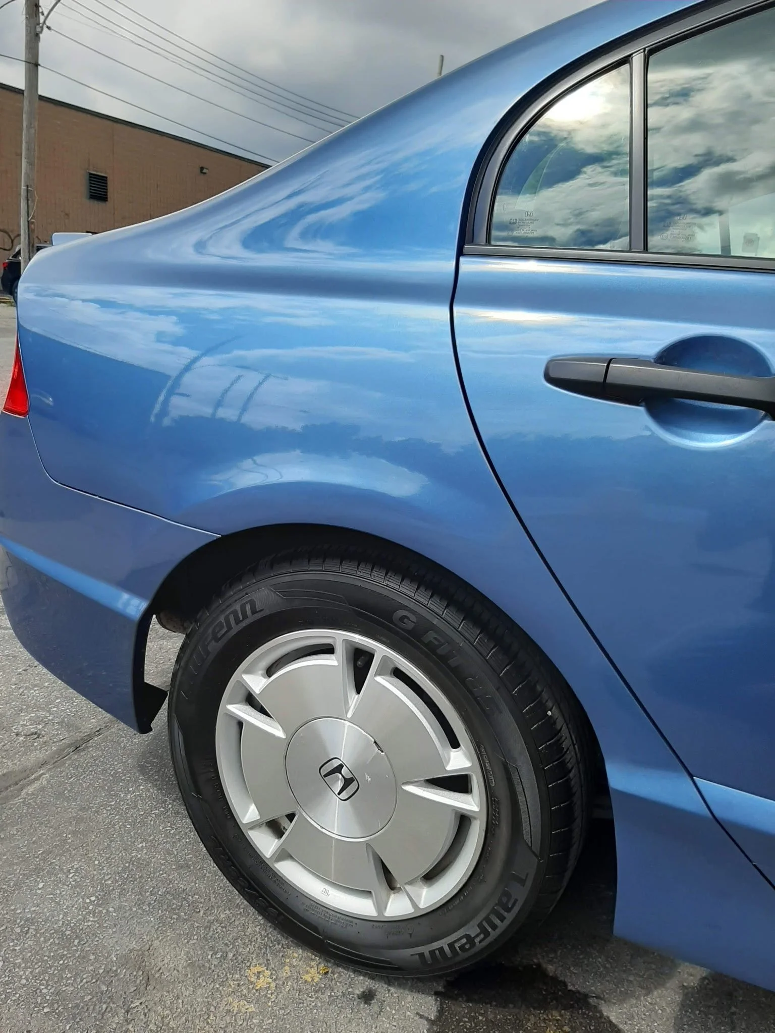 Close-up of a blue Honda sedan showing the rear wheel, part of the rear door, and the side window with reflections of clouds and sky.