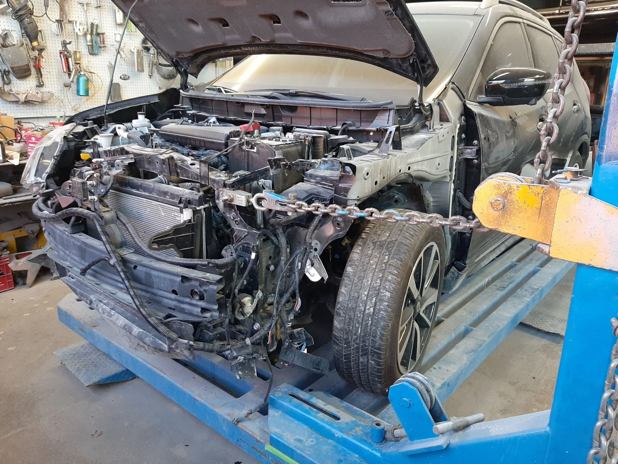 A black car with an exposed front engine and bumper, mounted on a blue repair lift, in a workshop with hanging tools and equipment, with part of the car's hood open and chained to the lift.