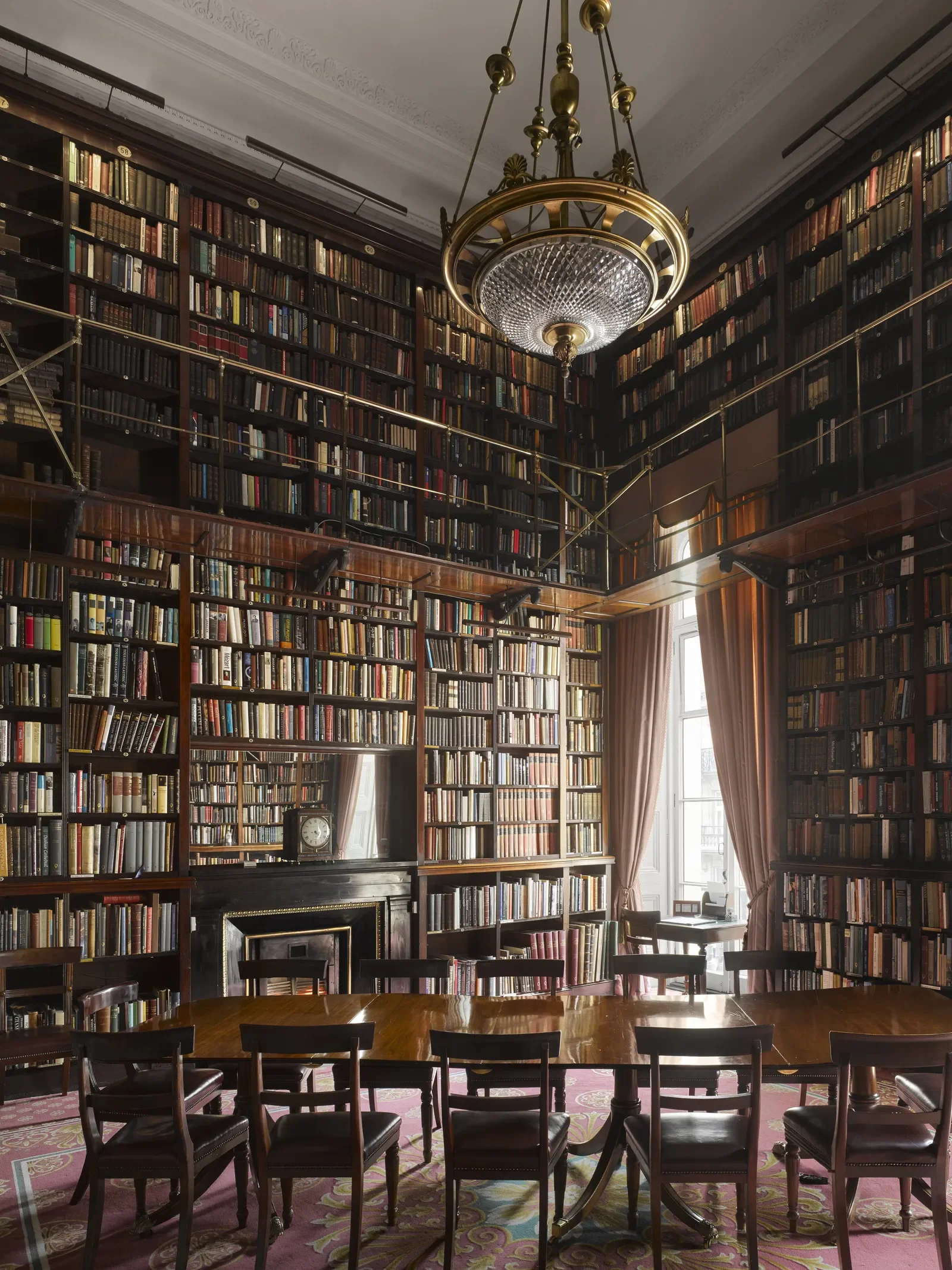 Two-story English library with floor-to-ceiling dark wood bookshelves, brass gallery ladder, antique crystal chandelier, and long wooden table at the Athenaeum Club London