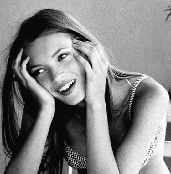 Black-and-white portrait of a young woman smiling with her hands at her face, wearing a patterned top in soft natural light.