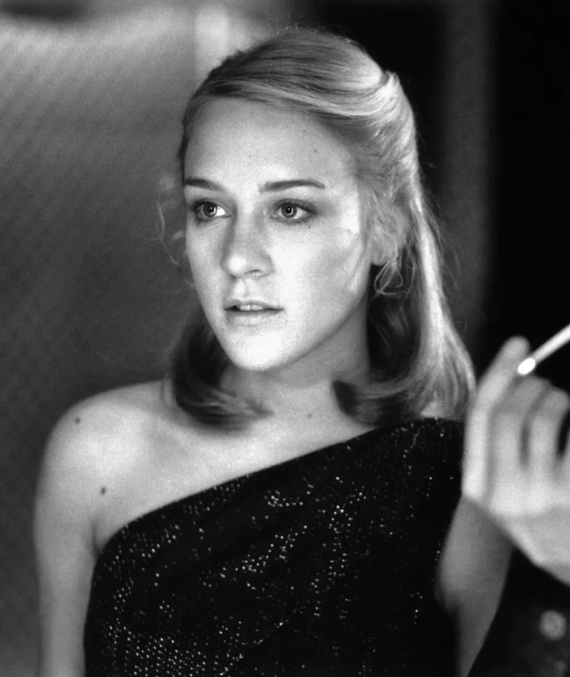 Black-and-white film still of a young woman in a one-shoulder sparkly dress, softly lit in a classic party scene.
