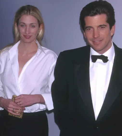 A man in a tuxedo and a woman in a white button-down shirt and black trousers standing together at an evening event, photographed against a plain backdrop.
