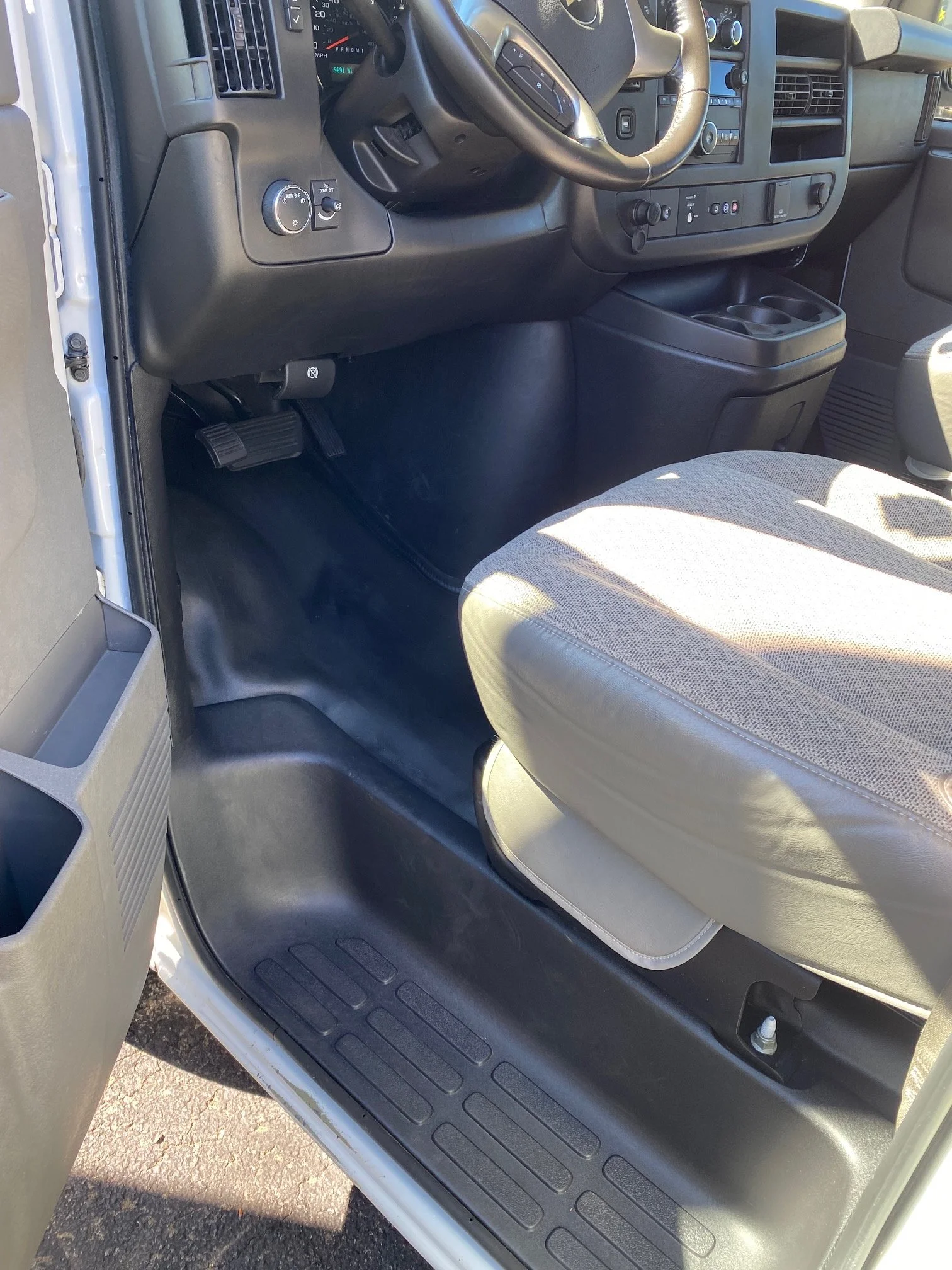 The image shows the interior of a vehicle, focusing on the driver's side foot area, the dashboard, and the steering wheel. It includes the accelerator and brake pedals, a section of the dashboard with control buttons, and a cup holder.
