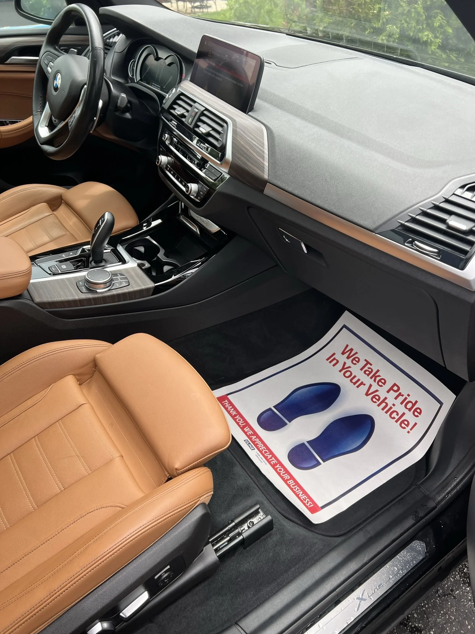 Interior of a BMW vehicle showing a beige leather seat, steering wheel, dashboard with a display screen, and a floor mat with red lettering that says "We Take Pride In Your Vehicle!".