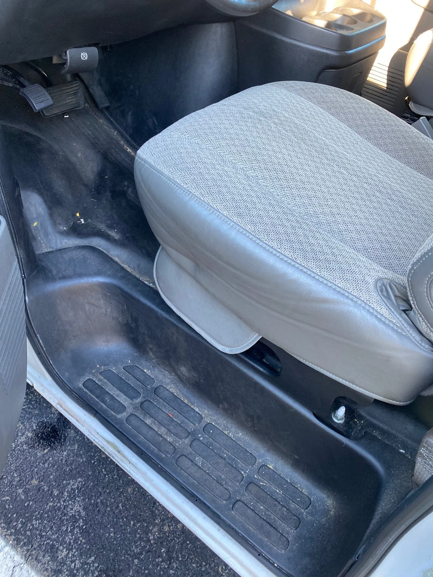 View of the passenger side floor area of a vehicle, including the footrest, brake pedal, and part of the seat with a fabric cover. The area appears slightly dirty with some small debris.