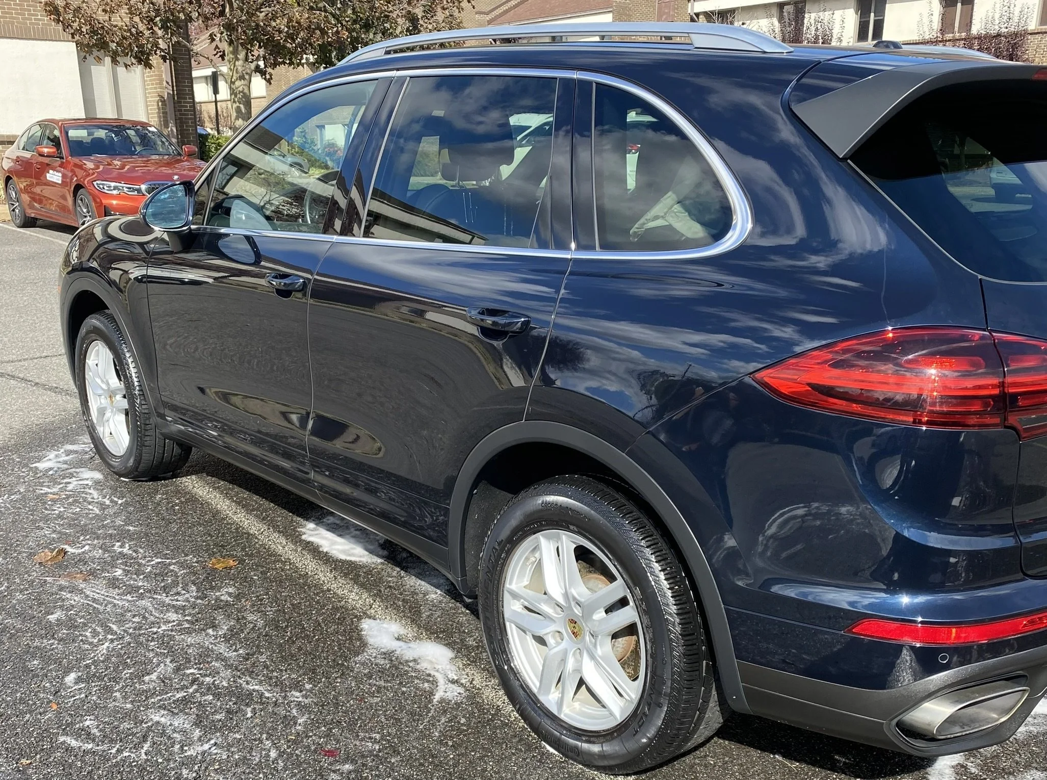 A black luxury SUV, possibly a Porsche, parked on a street with a red sedan parked in front of it. The scene is in a residential neighborhood with houses and trees nearby, and the ground is slightly snowy or icy.