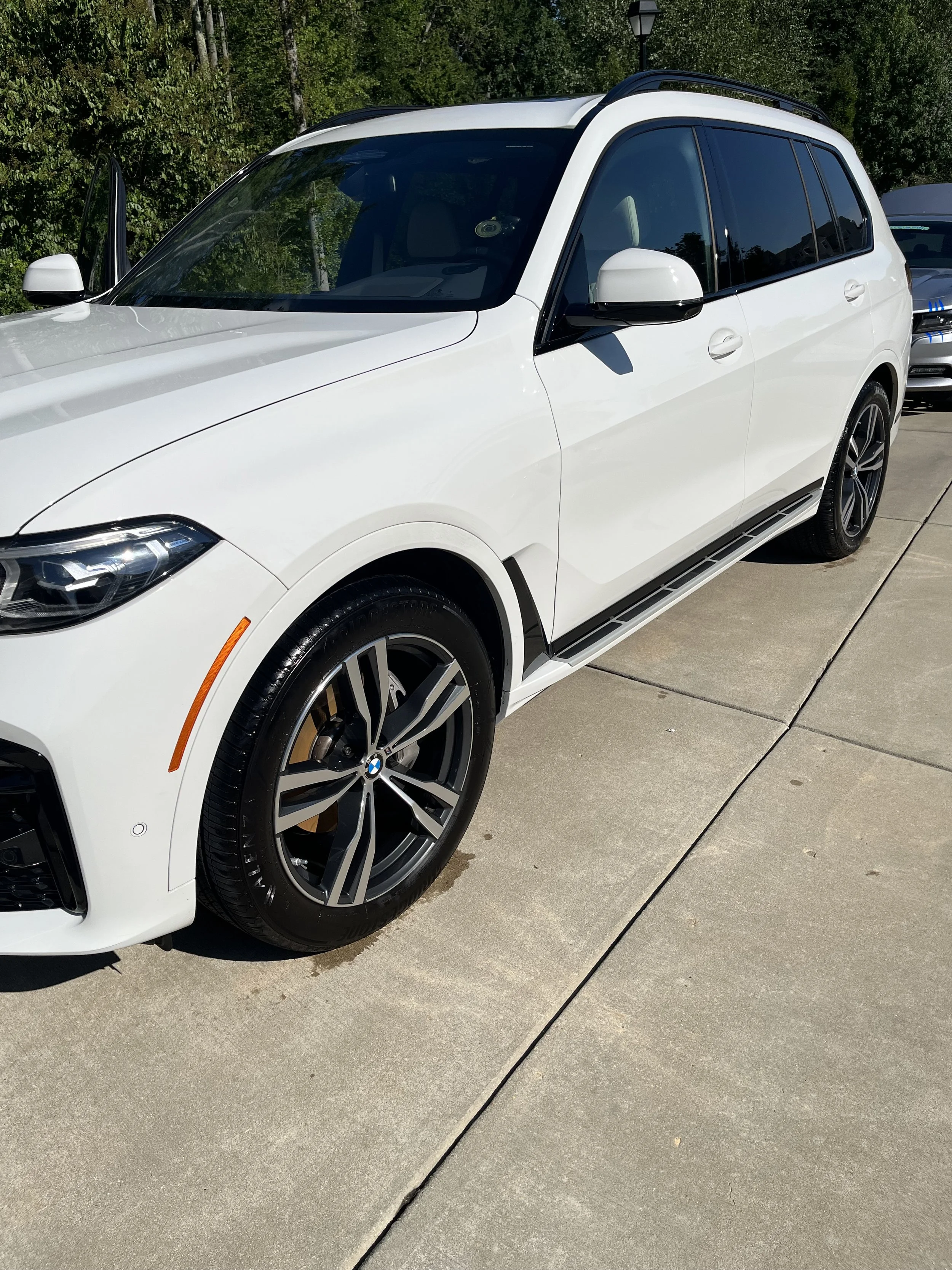 A white BMW SUV parked on a concrete driveway with green trees in the background and another vehicle partially visible behind it.