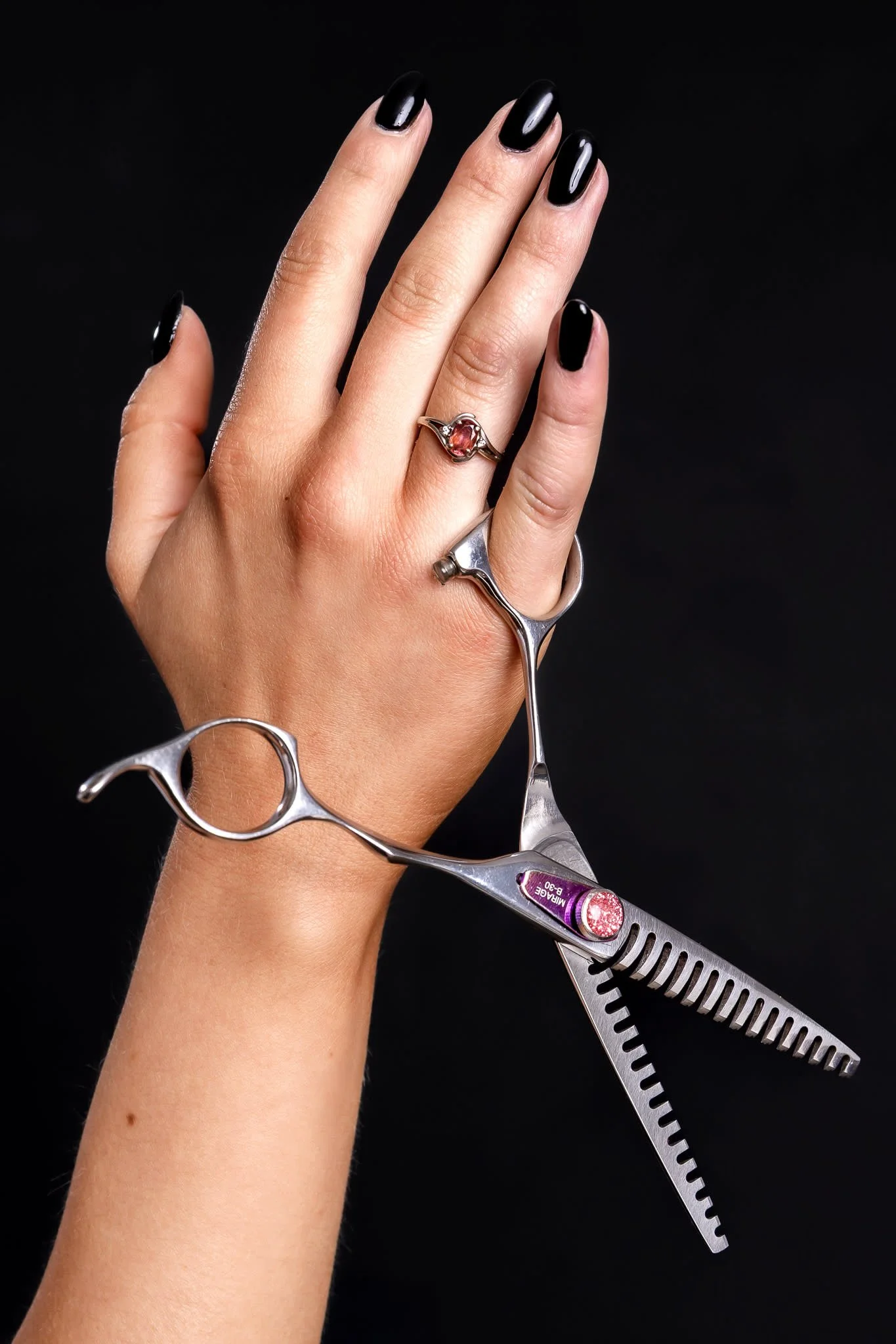 A hand with black nails holding hair cutting scissors and wearing rings, with a dark background.