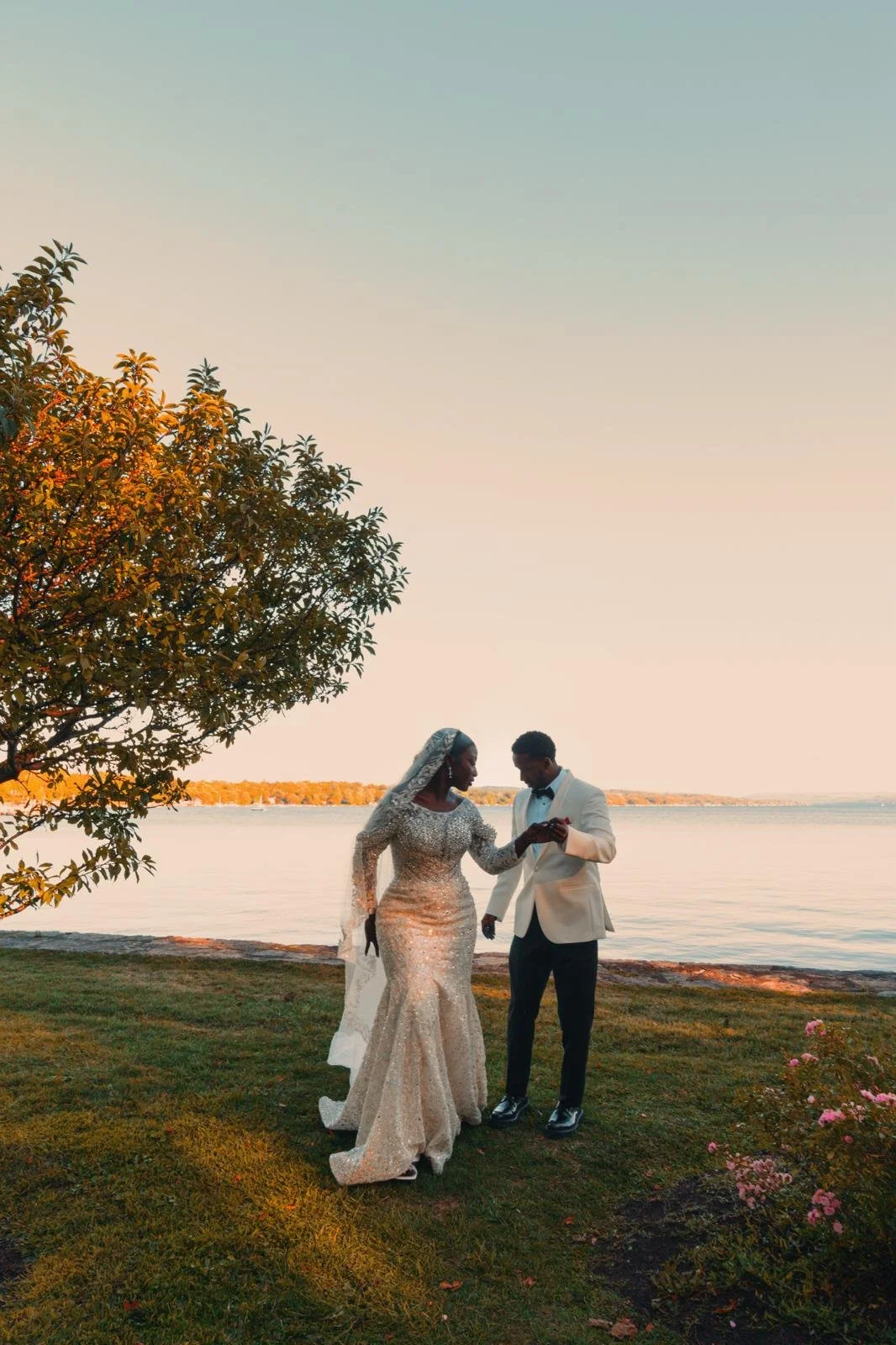 A bride and groom exchanging rings by a lakeside during sunset, with trees and flowers in the background.
