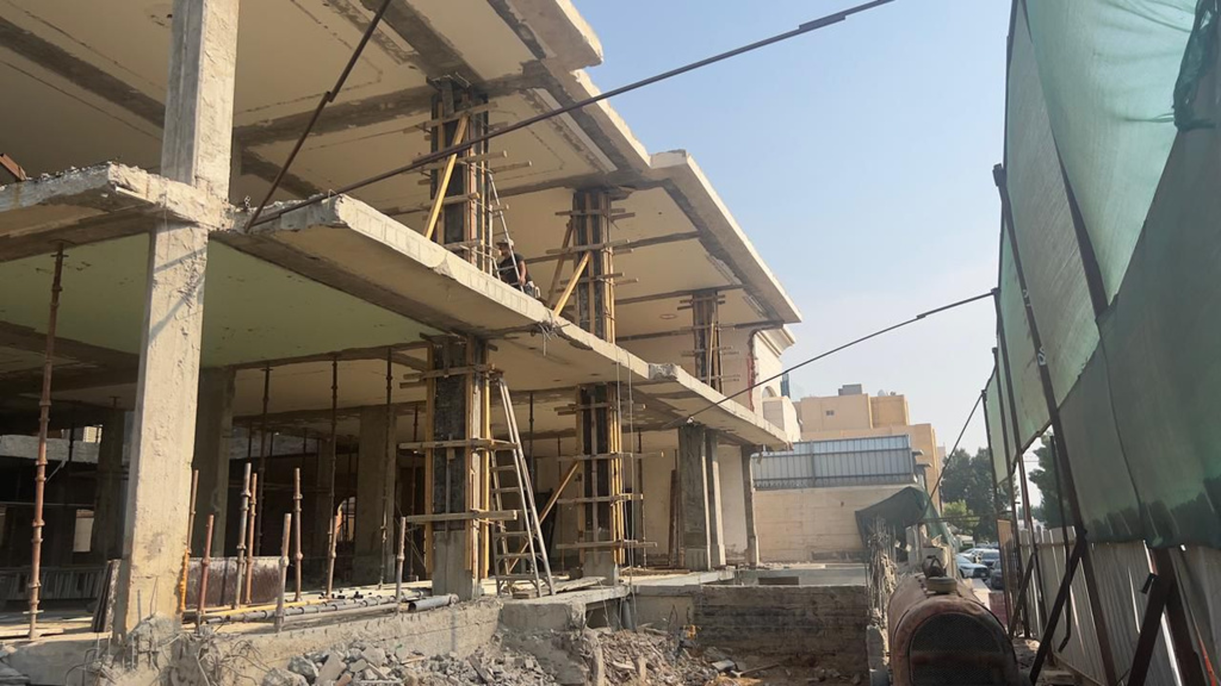 Construction site of a building with concrete floors and columns, scaffolding, ladders, and construction workers. Green construction tarp on the right side, with a vehicle and parked cars in the background.