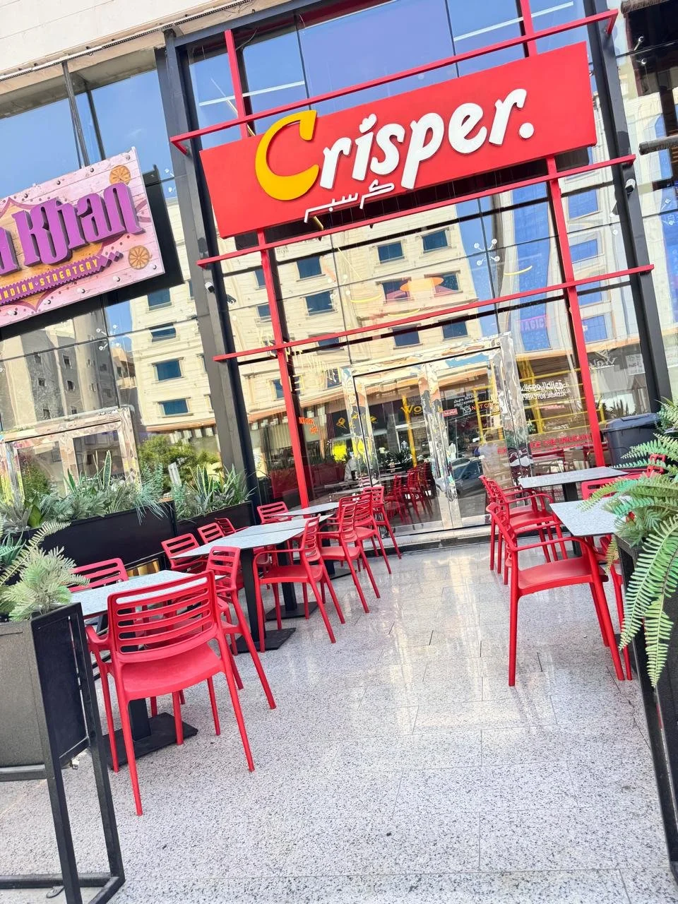 Outdoor seating area of Crisper restaurant with red chairs and tables in front of a glass facade, reflected buildings, and surrounding planters.