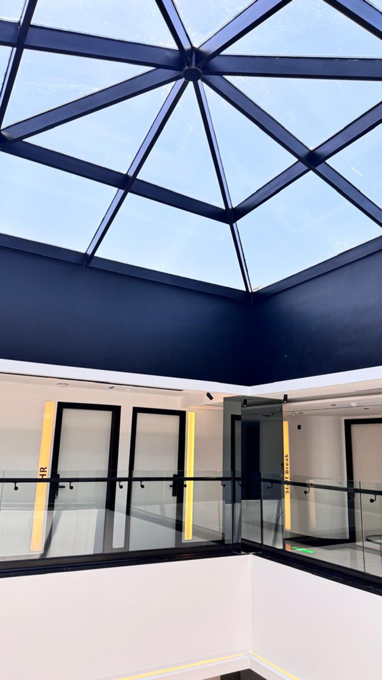 Interior of a modern building with a glass ceiling and black railings, including exit signs labeled 'HR' and 'Staff Break'.