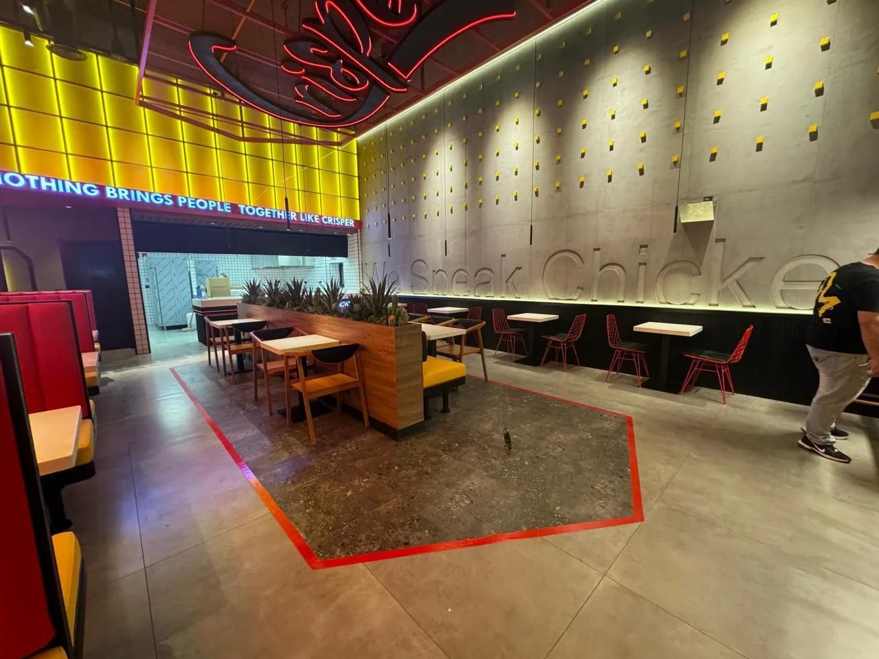Interior of a modern restaurant with colorful neon lights, yellow accent walls, and various seating options including booths and individual tables, with a concrete wall featuring the words 'speak, ch lick' carved into it.