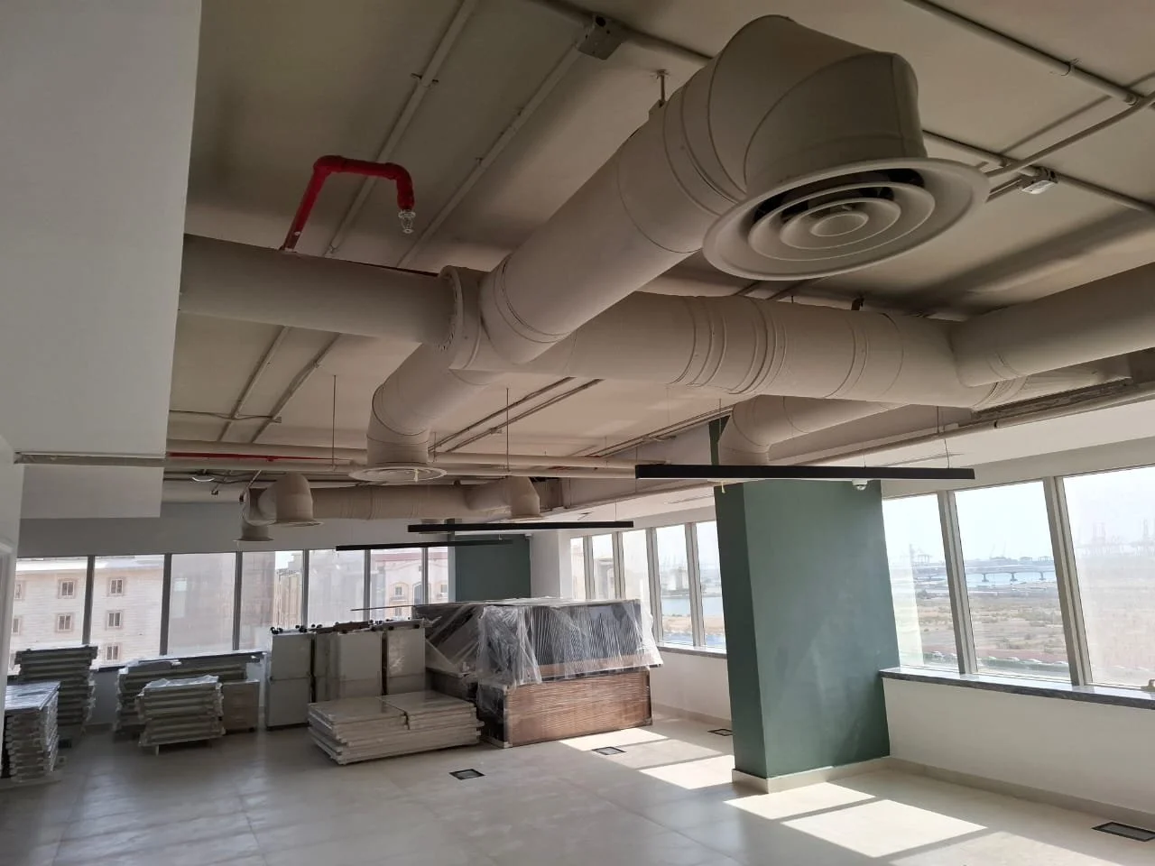 An office or commercial space under construction with exposed ceiling pipes and ductwork, stacks of building materials, and large windows with a view outside.