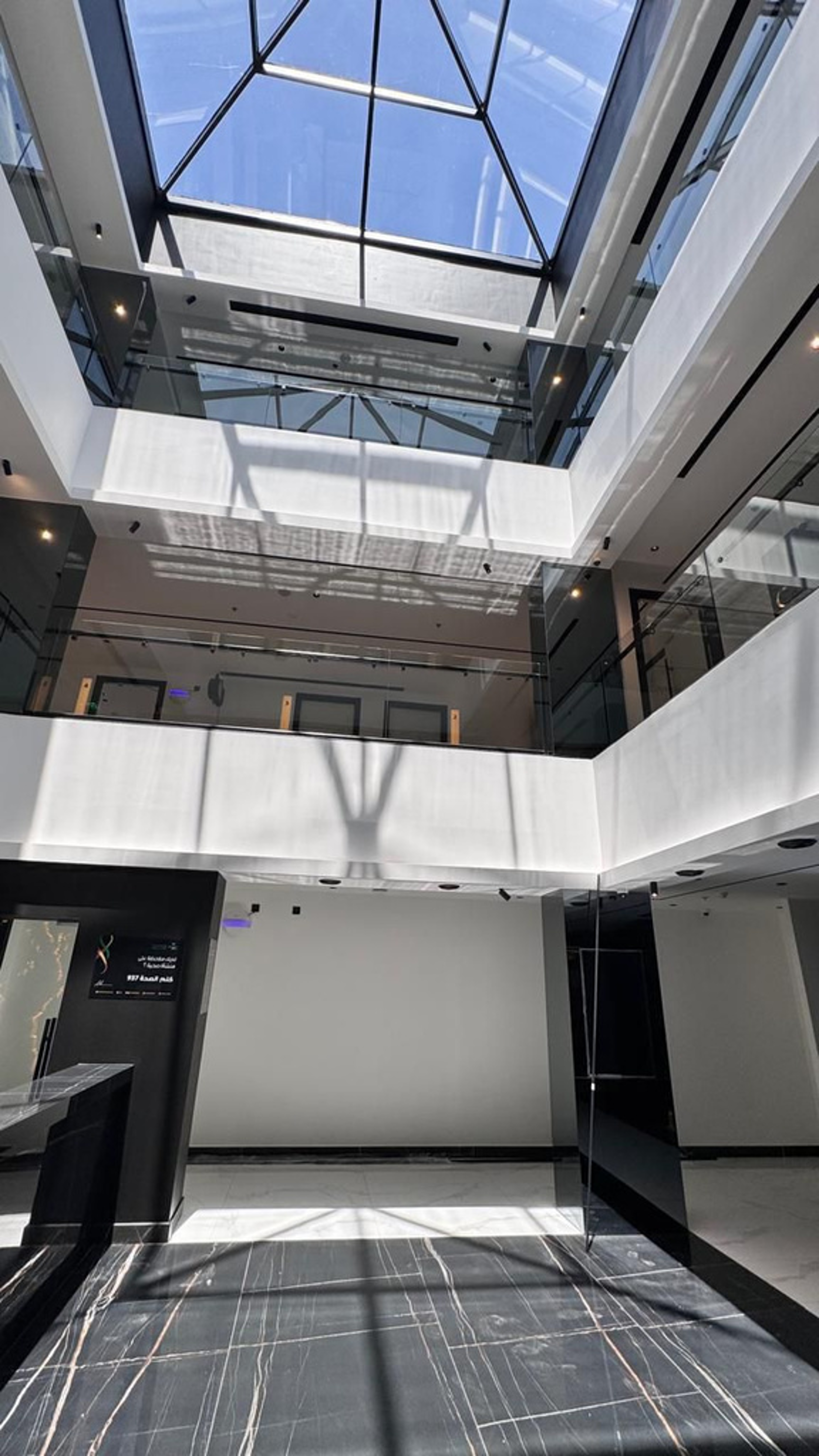 Interior view of a modern building with a large skylight at the top, white walls, black marble flooring with white veins, glass railings, and multiple floors visible.