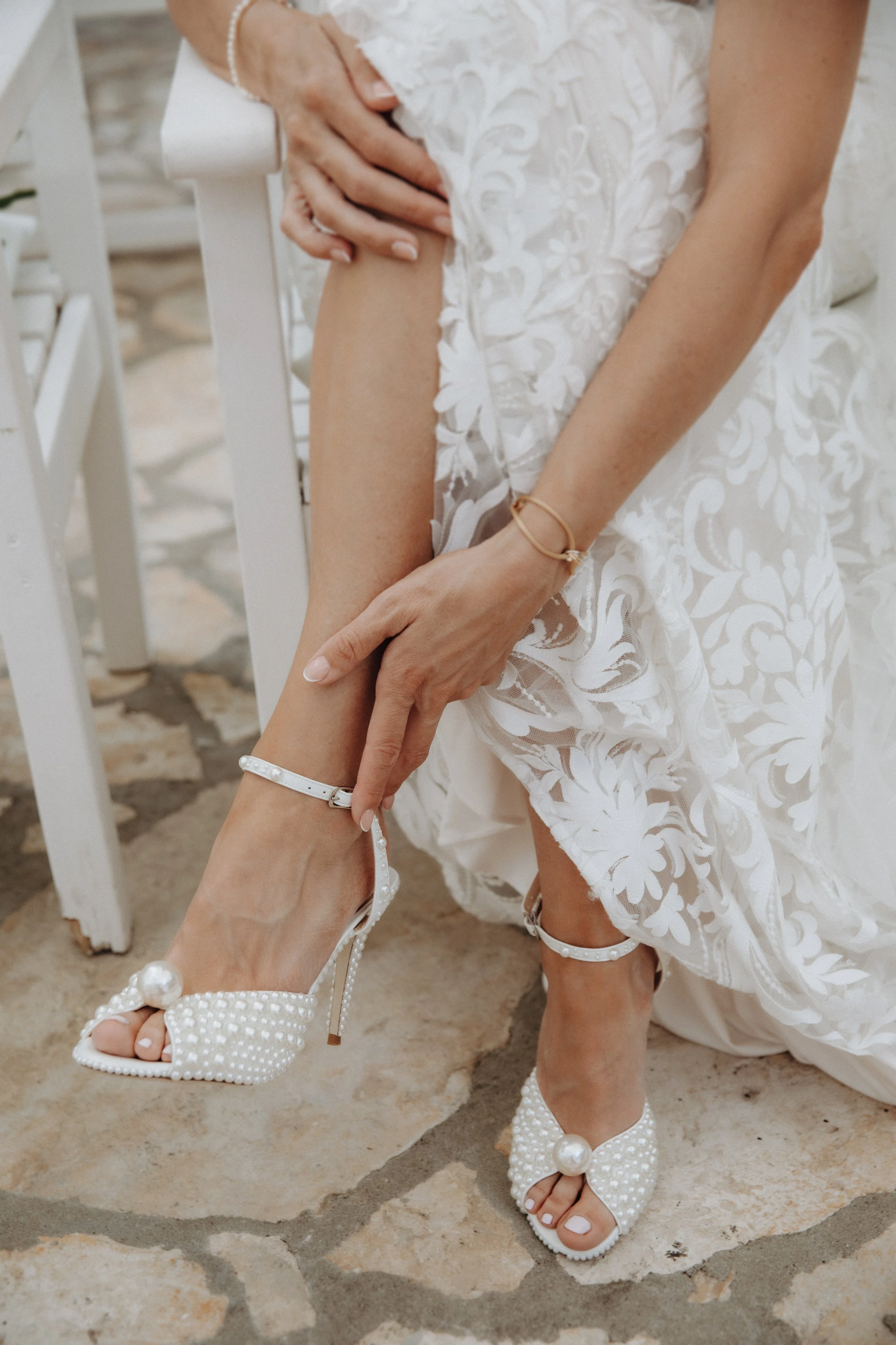 Eine Frau in einem weißen Hochzeitskleid zieht auf einer Steinterrasse ihre weißen Perlen-High-Heels an.