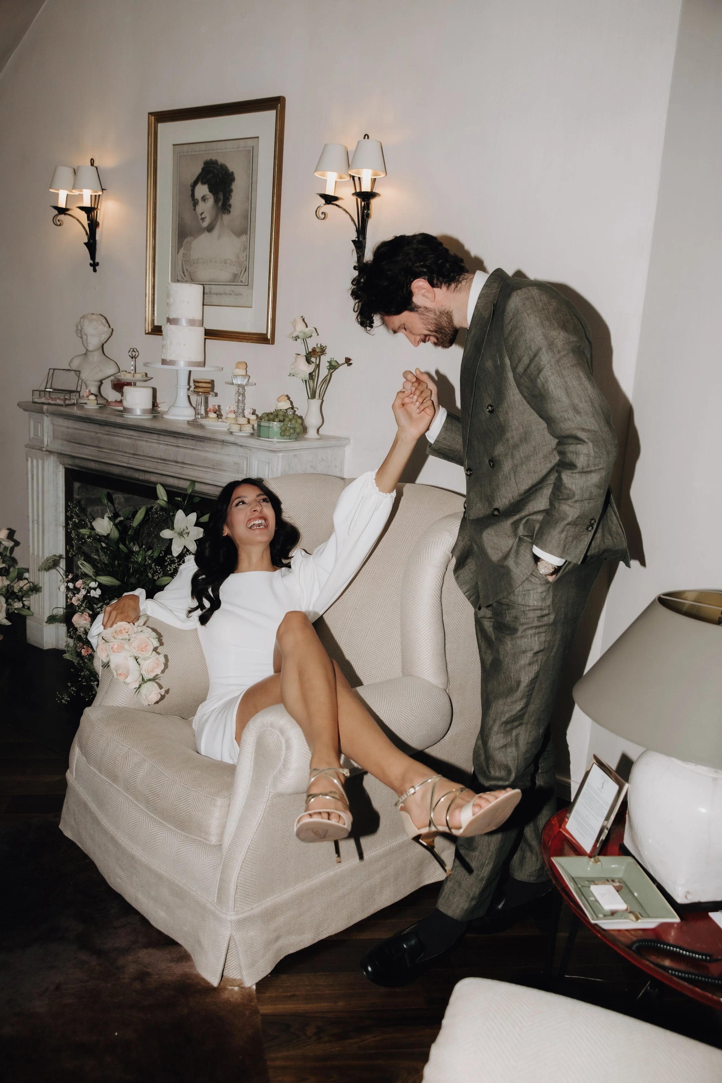 Ein Mann und eine Frau tanzen und lachen in einem Wohnzimmer. Die Frau sitzt auf einem Sofa, die Frau trägt ein weißes Kleid und high heels. Der Mann trägt einen grauen Anzug.