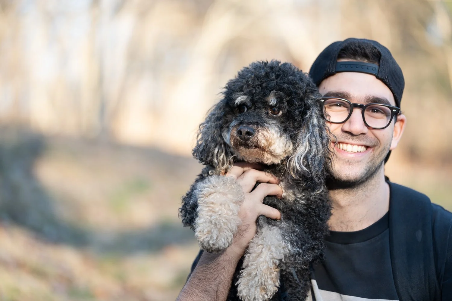 Man in backwards hat and glasses smiling holding bernedoodle dog.