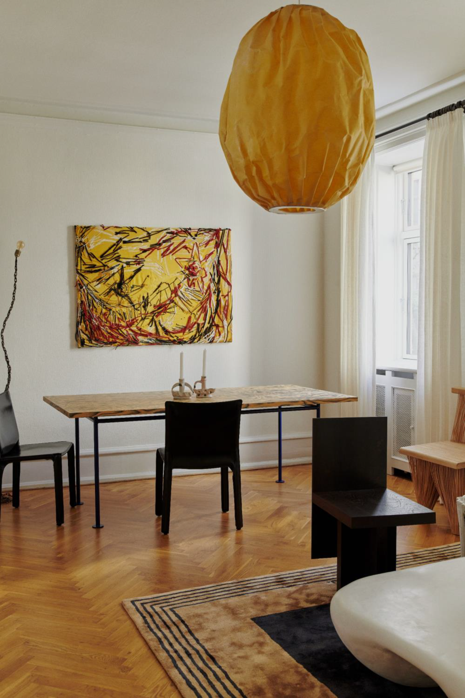 A room with a wooden dining table, black chairs, abstract wall art, a paper lantern hanging from the ceiling, and a window with white curtains. There is a round white chair and a black table in the foreground on a hardwood floor.