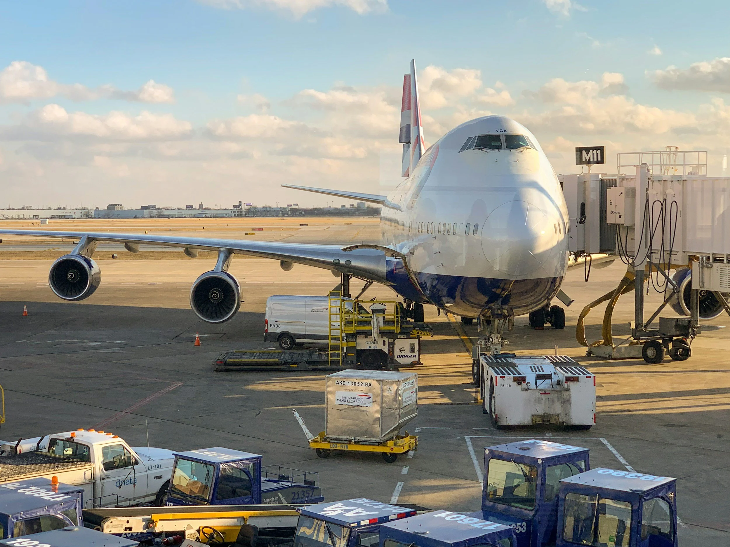 A large commercial airplane parked at an airport gate with jet bridges and ground service vehicles around it, under a partly cloudy sky.