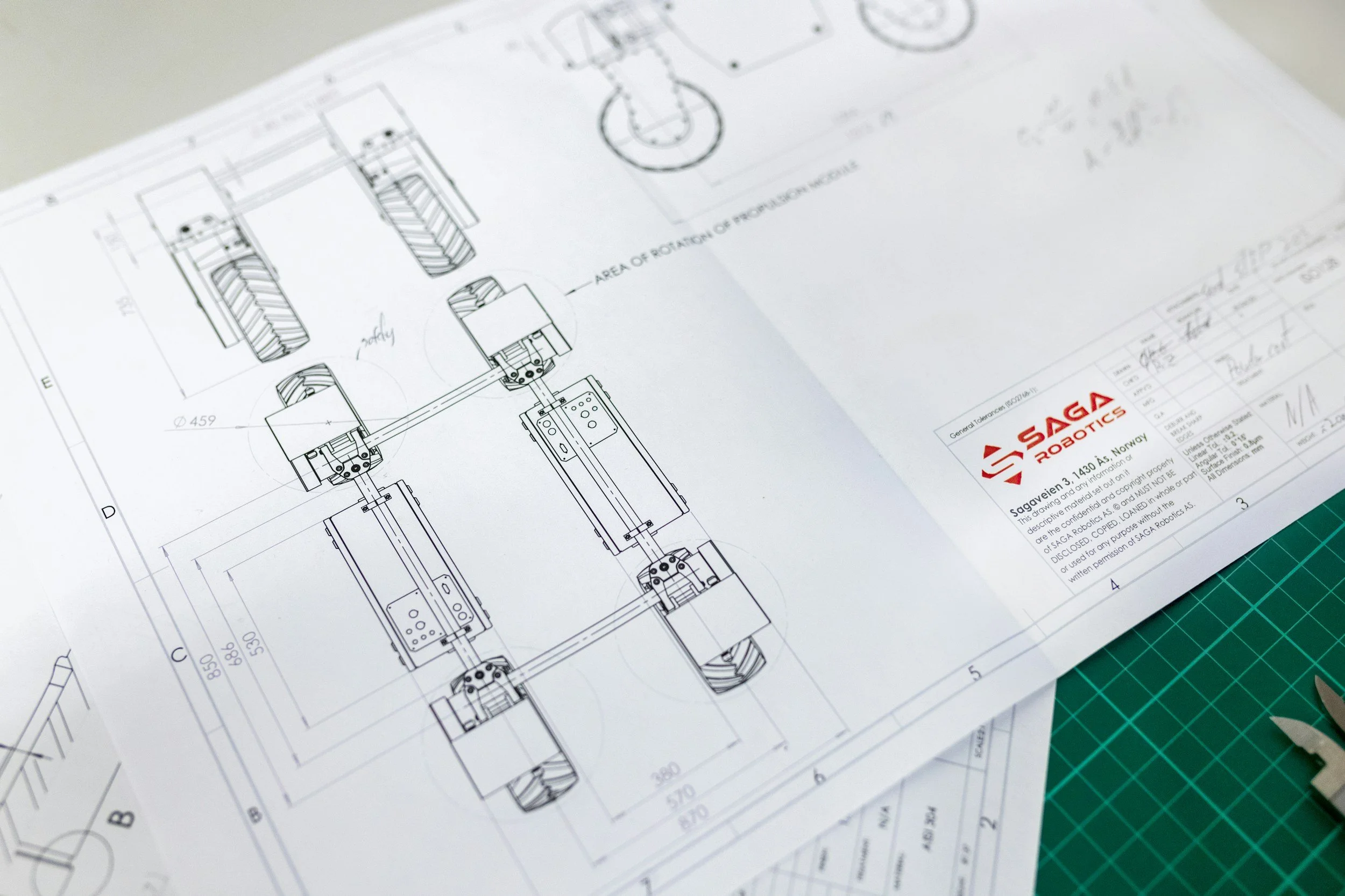 Technical engineering drawing on white paper, featuring mechanical parts and measurements, with a green cutting mat underneath.