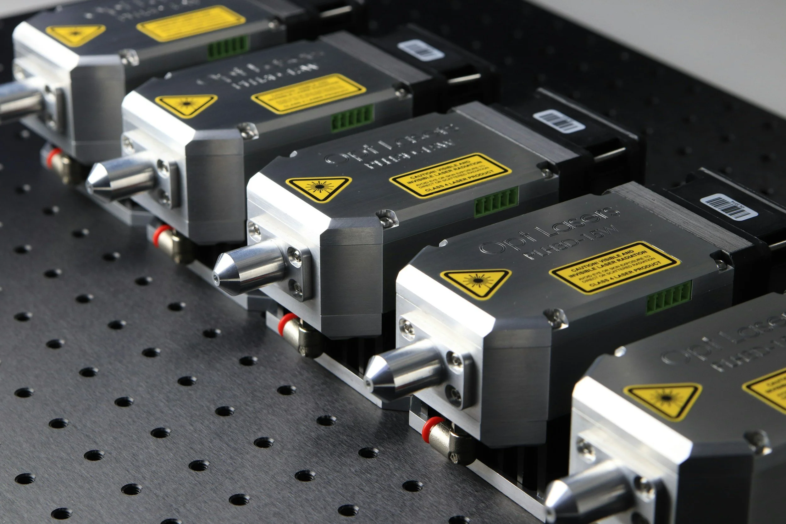 Three laser modules mounted on a perforated metal surface, each with yellow warning labels and warning symbols, aligned in a row for precision laser applications.