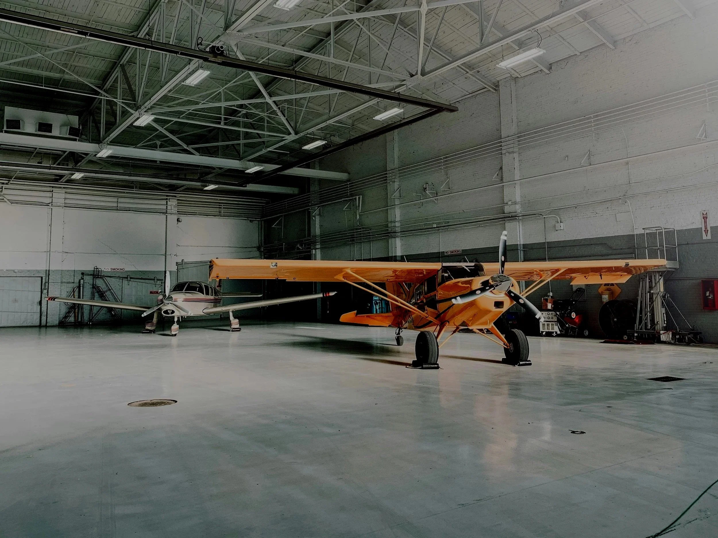 The image shows the interior of an aircraft hangar with a yellow vintage airplane parked on the floor. The hangar has a high ceiling with exposed metal beams and ample lighting. The walls are made of white bricks, and there's some equipment and a fire extinguisher visible in the background.