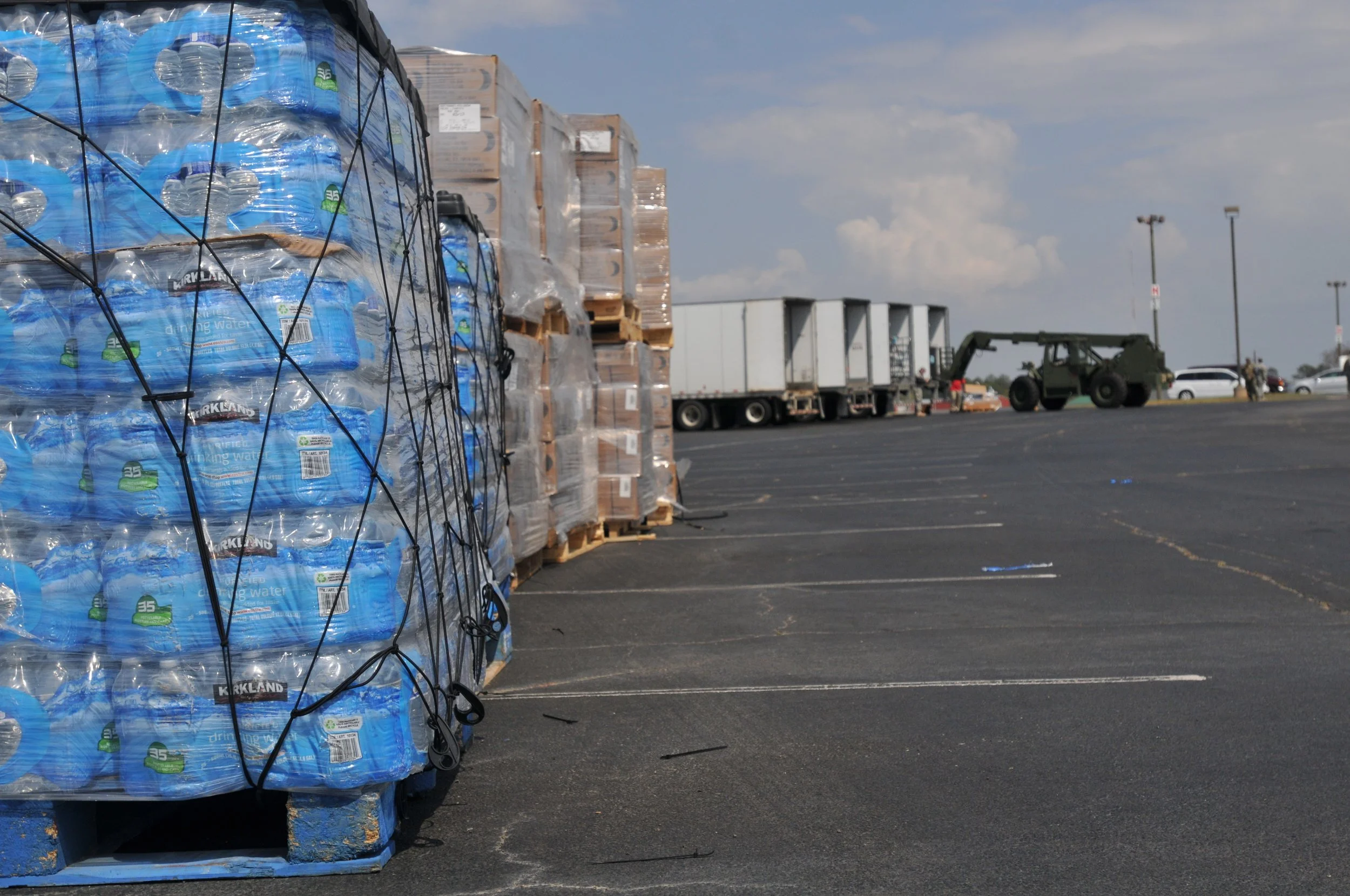 Stacked pallets with bottled water wrapped in plastic on a parking lot, with trucks and cars in the background under cloudy skies.