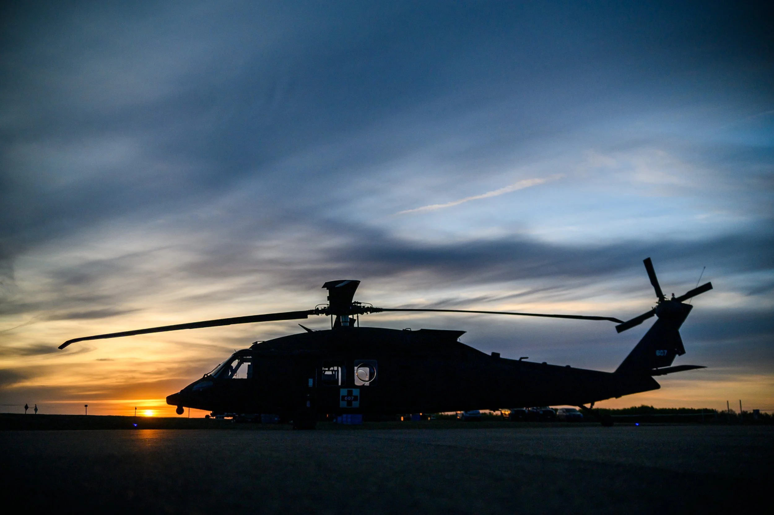 Silhouette of a military helicopter parked outdoors during sunset or sunrise with a darkening sky and clouds in the background.