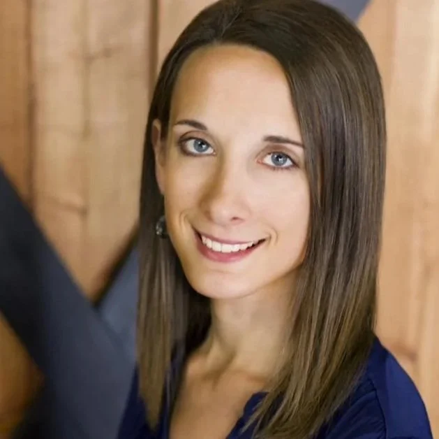 A young woman with long brown hair and blue eyes, smiling and looking at the camera, with a wooden background.