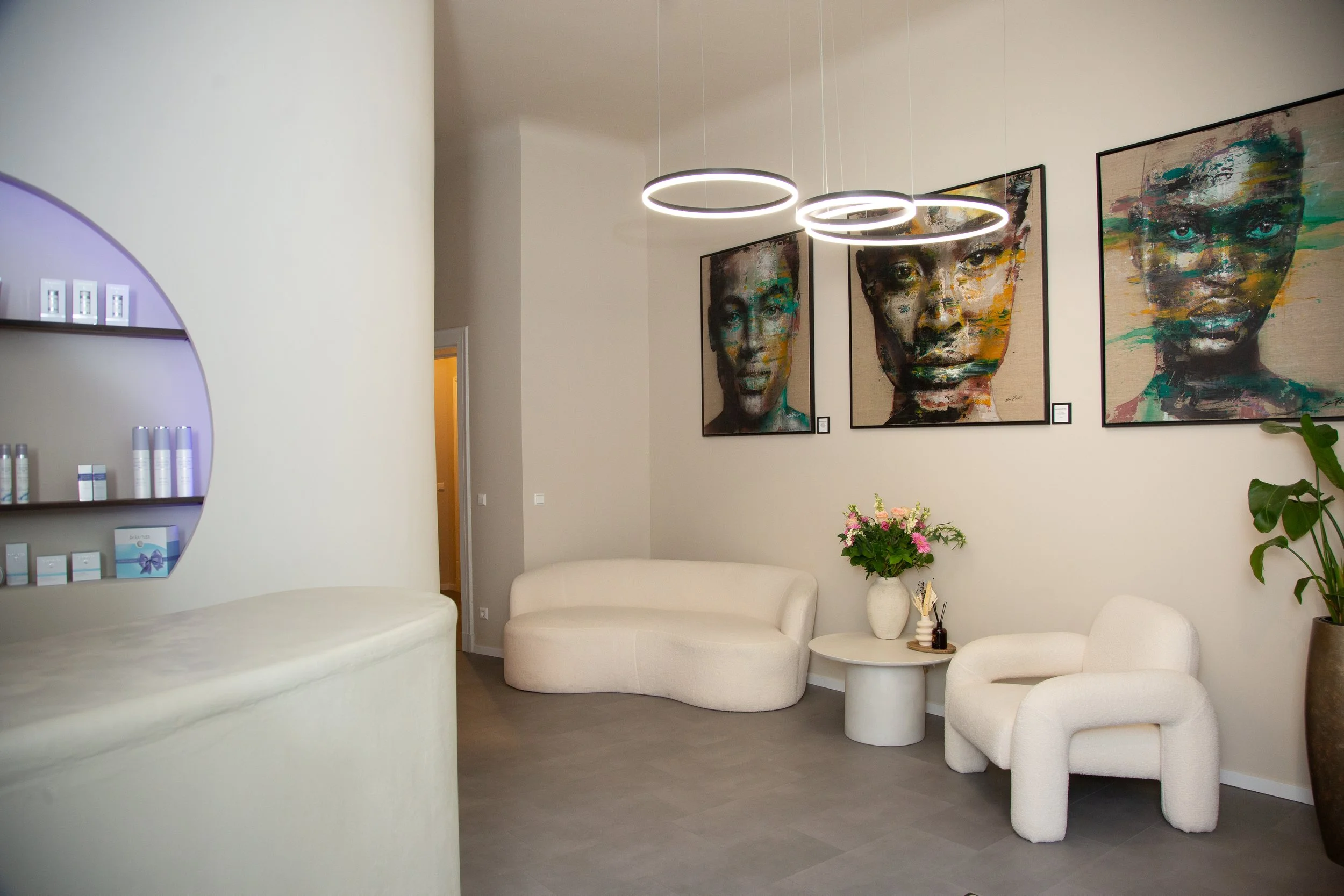 Gemütlicher Raum mit weißen Sesseln, einem kleinen runden Tisch mit Blumen, modernen Wandkunstporträts und dekorativen Lichtringen.