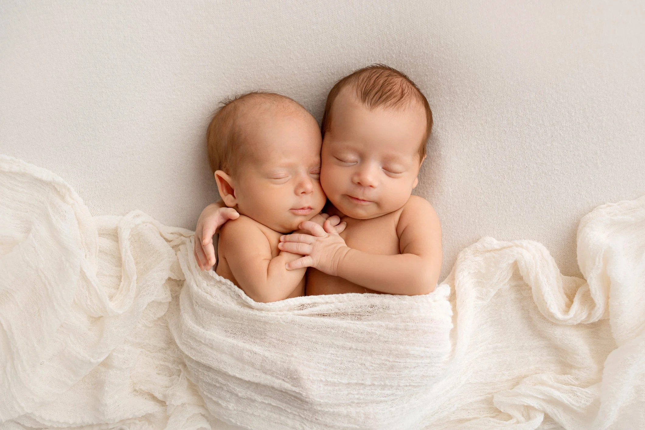 Two sleeping babies, wrapped in a soft white blanket, cuddling closely against a beige background.
