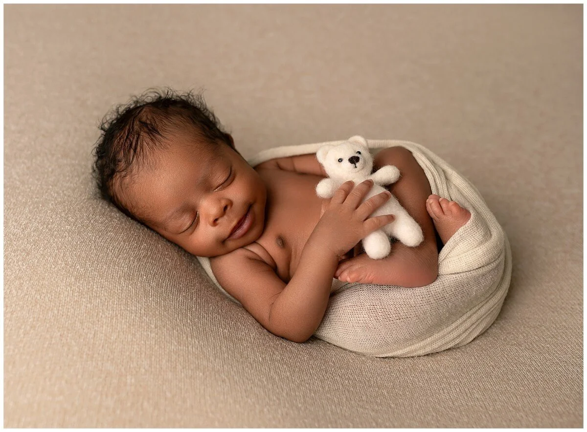 Baby sleeping on a beige surface, holding a small white stuffed bear.