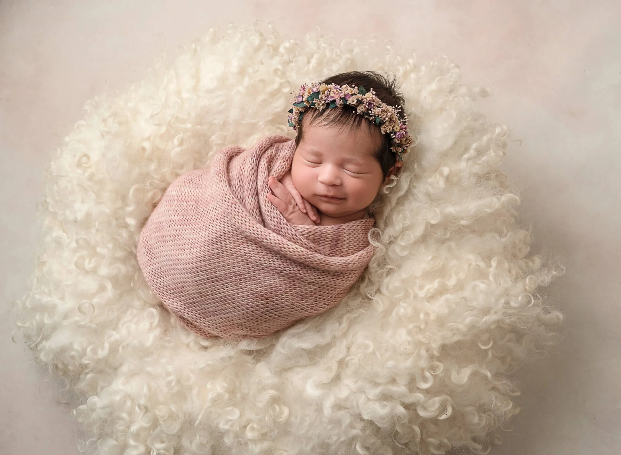 A newborn baby wrapped in a pink knit blanket, wearing a floral headband, sleeping on a fluffy, curly white blanket.