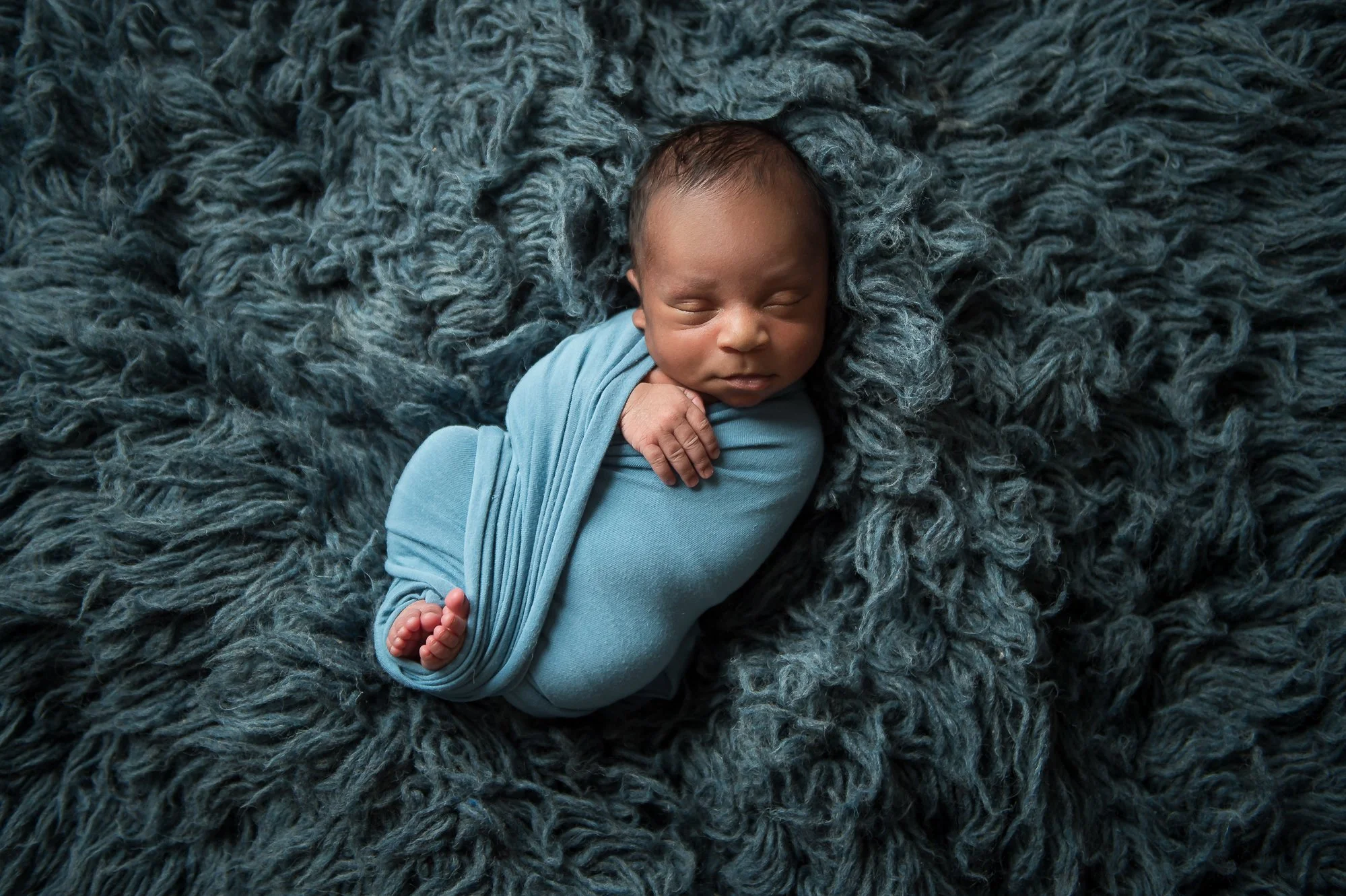 A newborn baby sleeping on a fluffy, textured dark blue blanket, wrapped in a matching light blue cloth.