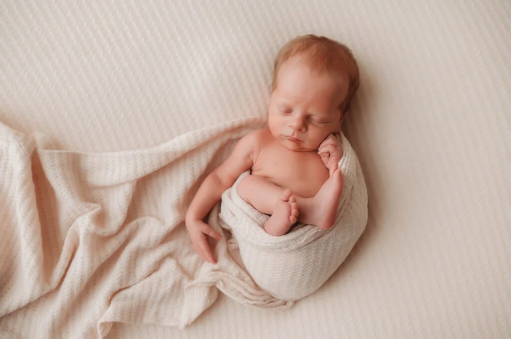 Newborn baby sleeping, wrapped in a beige blanket, on a cream-colored textured surface.